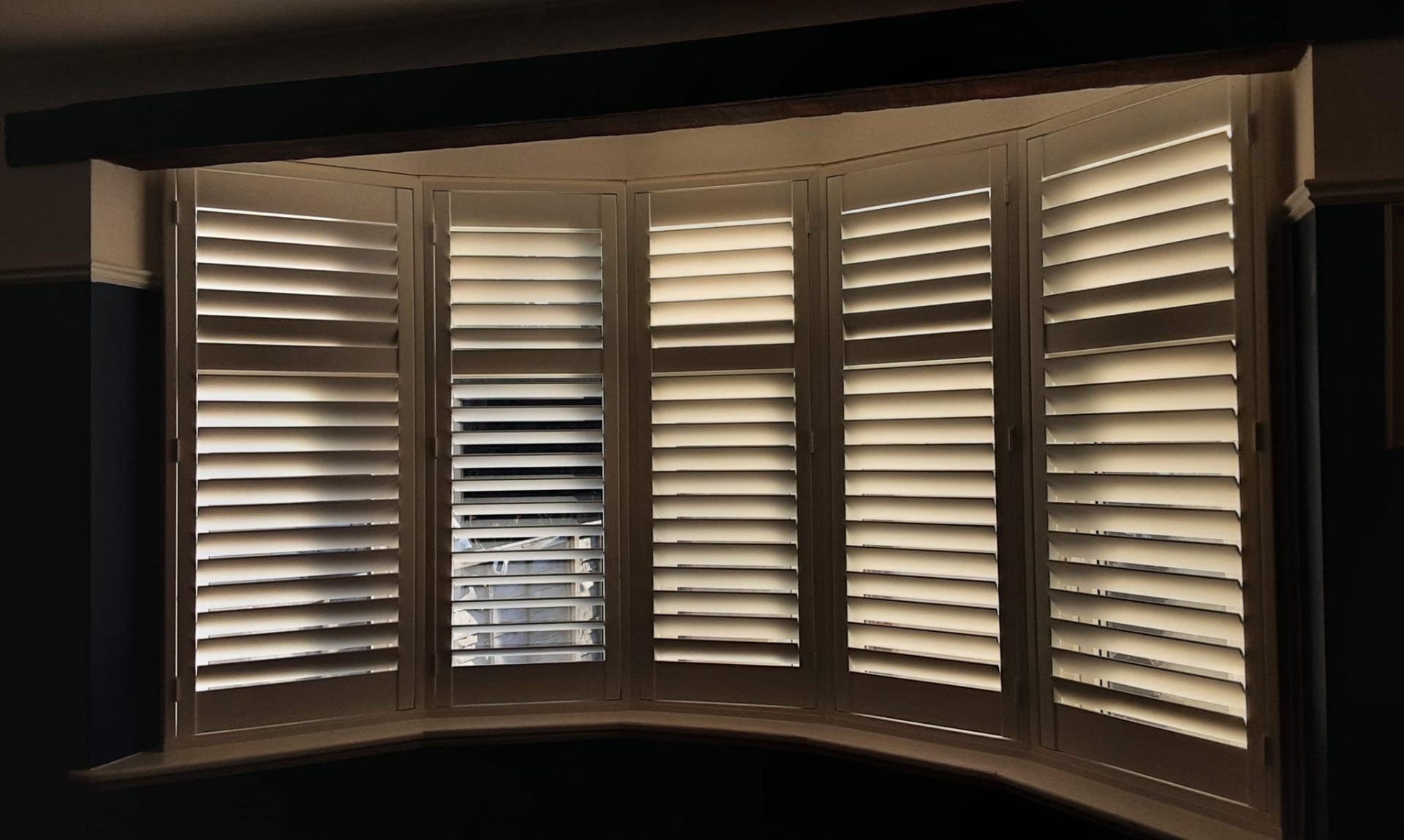 Bay window plantation shutters with adjustable louvres in a dark-toned room