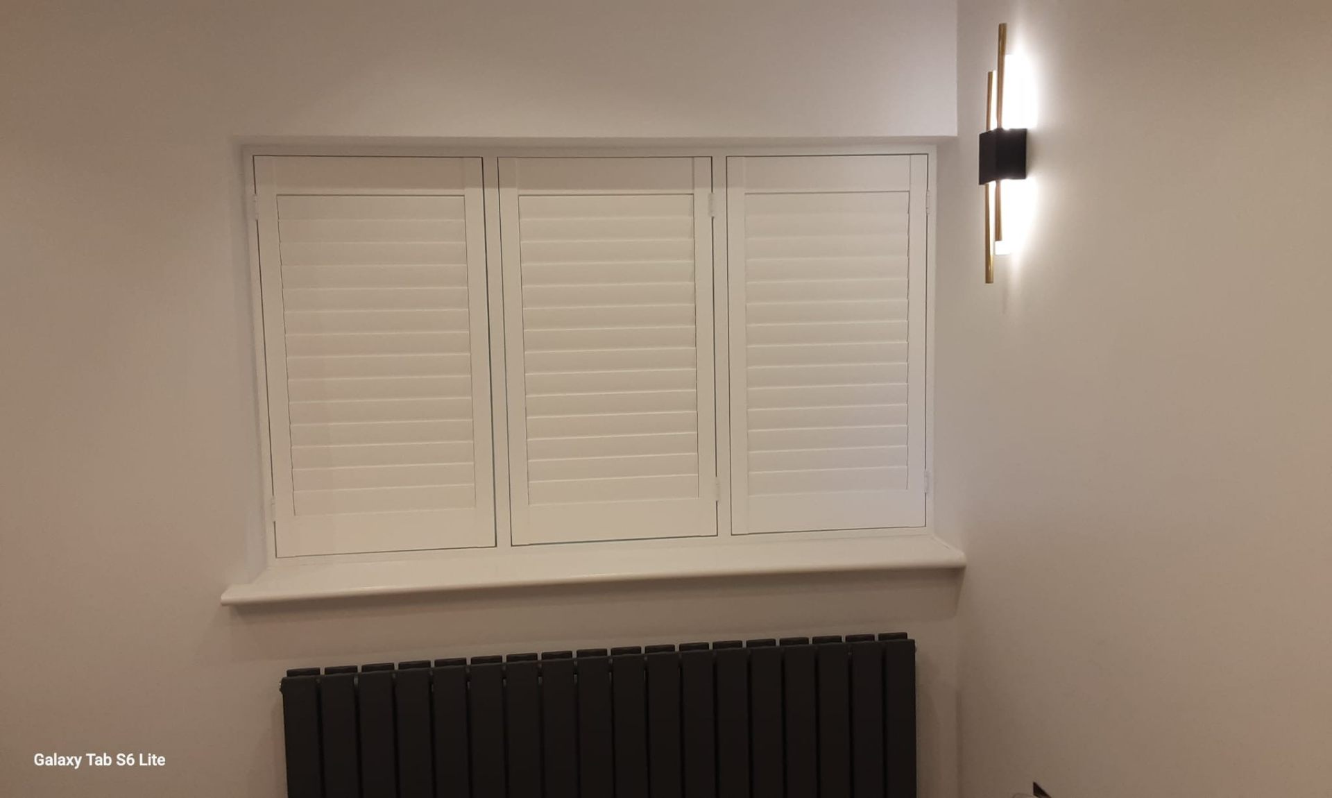 Three-panel plantation shutters closed above radiator with wall light
